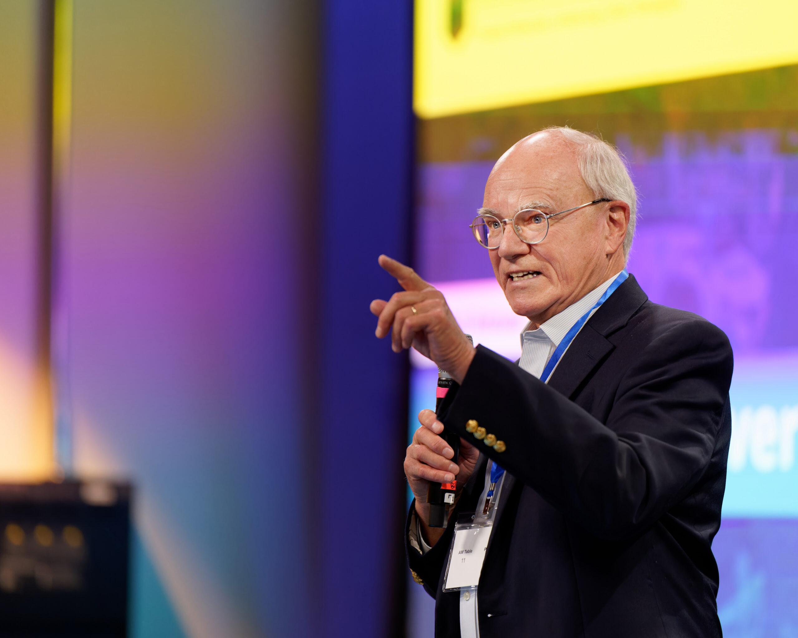 Conference speaker holding a microphone and gesturing on a purple-and-blue stage